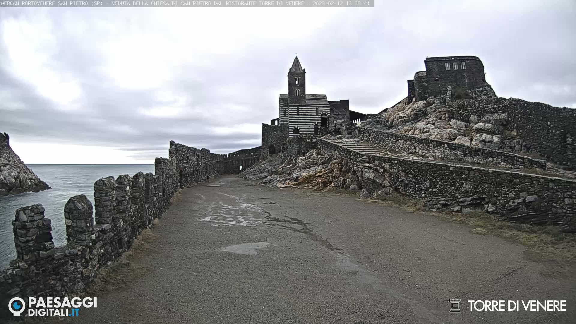 A stone path leads to a striped church and stone structures atop a rocky cliff overlooking a calm sea under a cloudy sky.