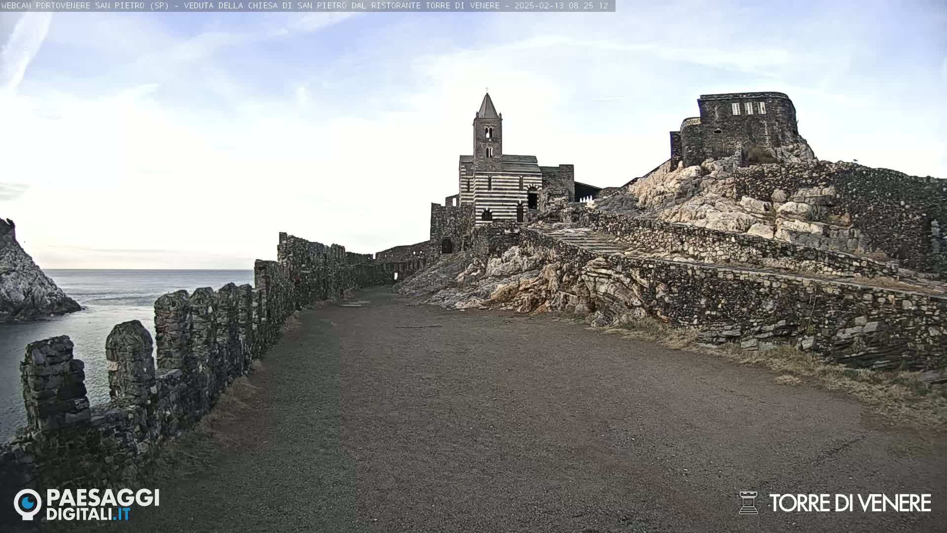 A stone church and ruins are situated on a rocky cliff overlooking a calm sea under a mostly clear sky.