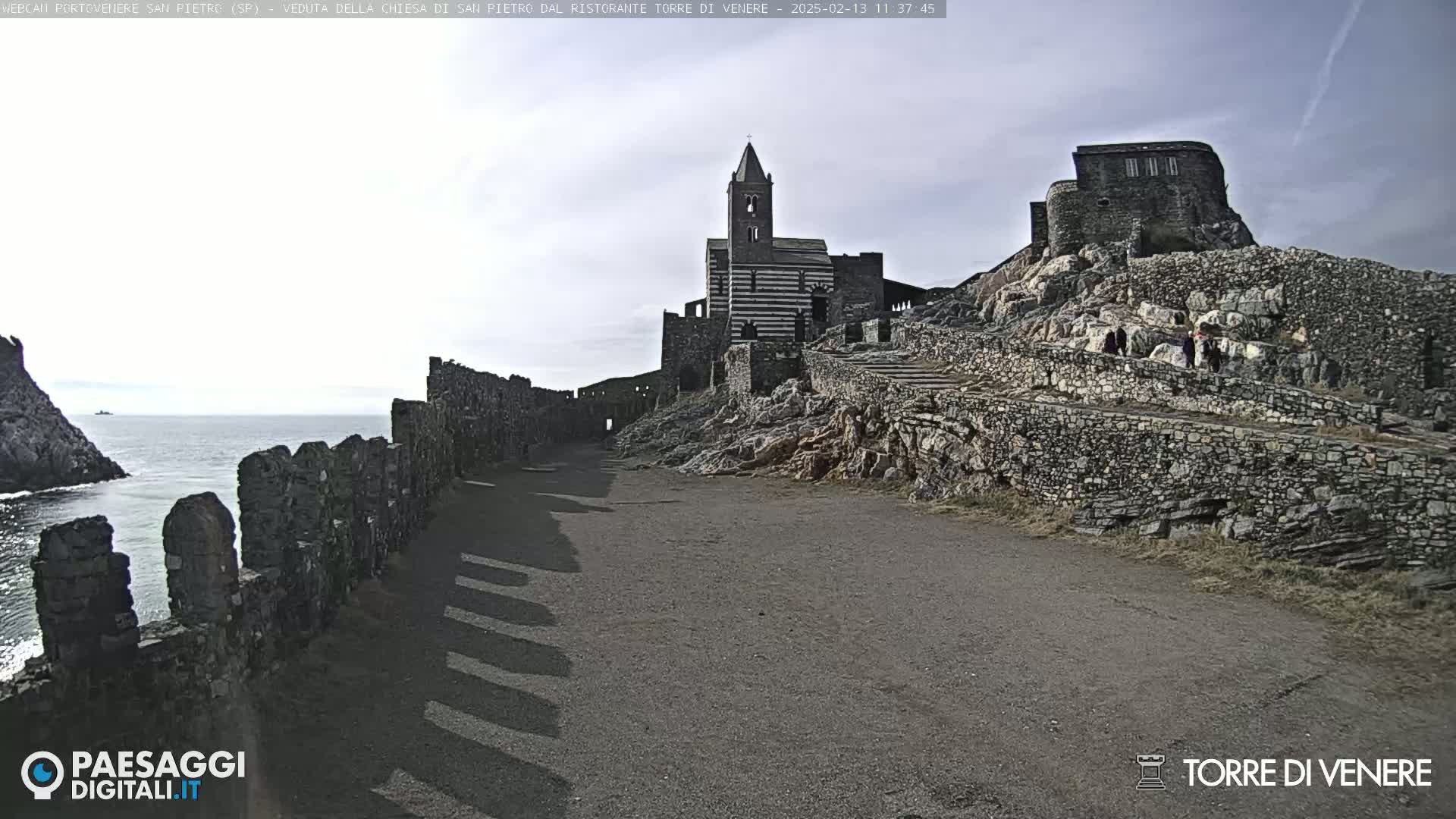 A stone pathway leads to a striped church and ruins on a rocky hillside overlooking a calm ocean under a mostly cloudy sky.