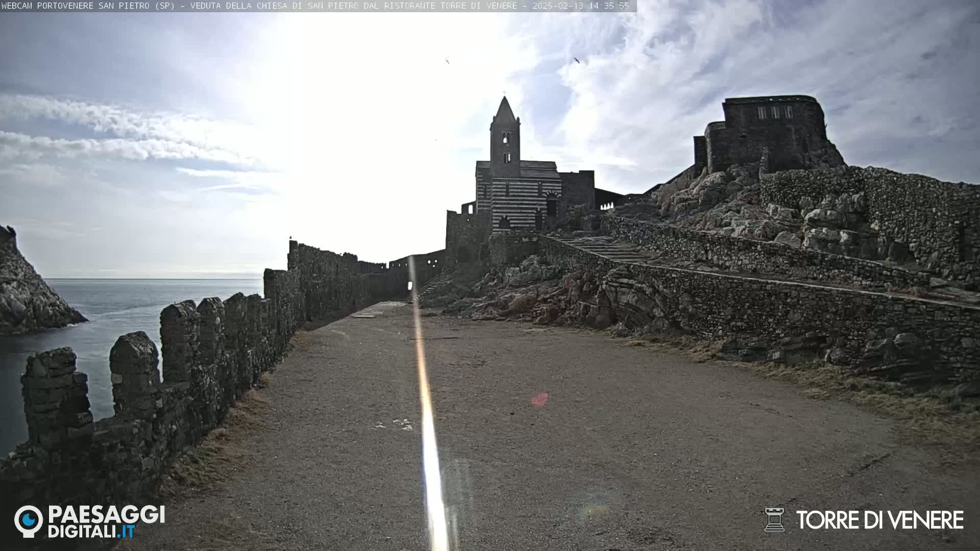 A stone path leads to a striped church and castle ruins perched on a rocky cliff overlooking a calm sea under a partly cloudy sky.