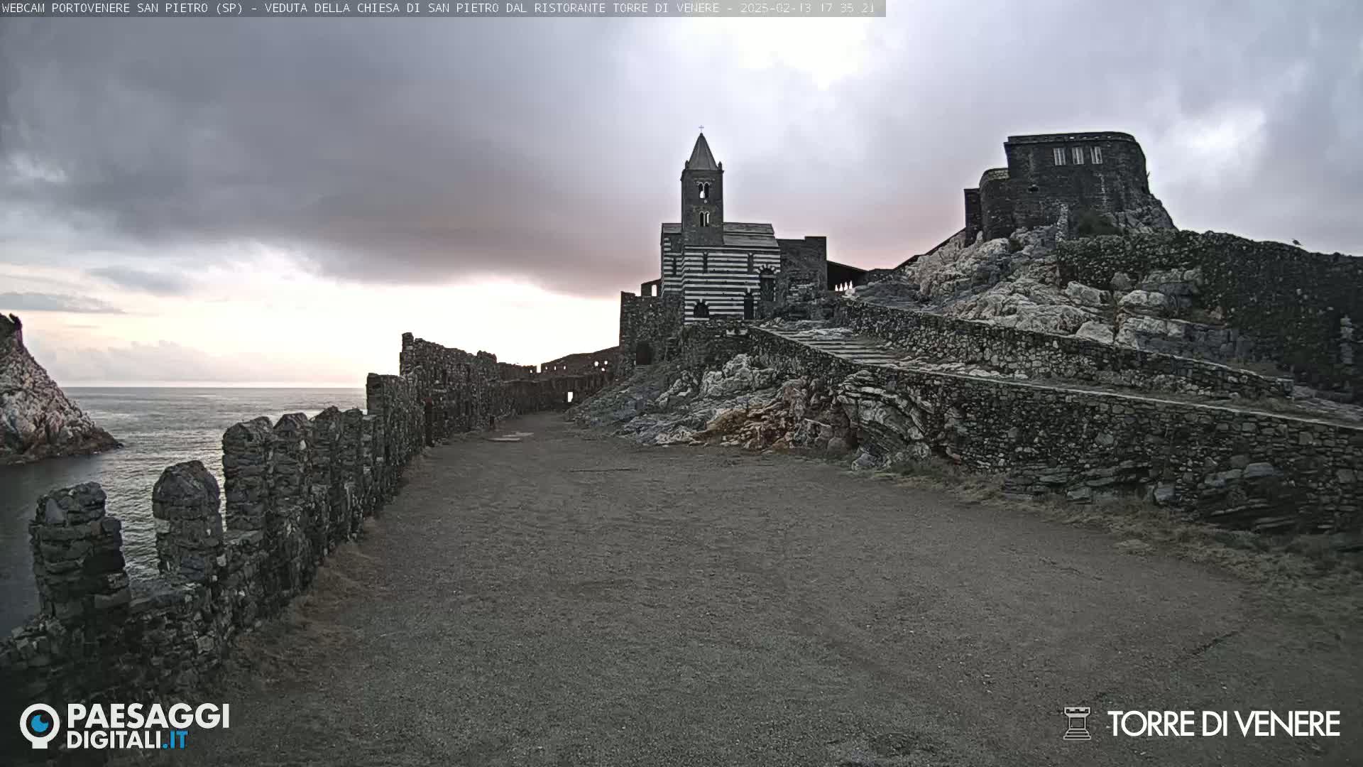 A stone church and ruins sit atop a rocky cliff overlooking a calm sea under an overcast sky.