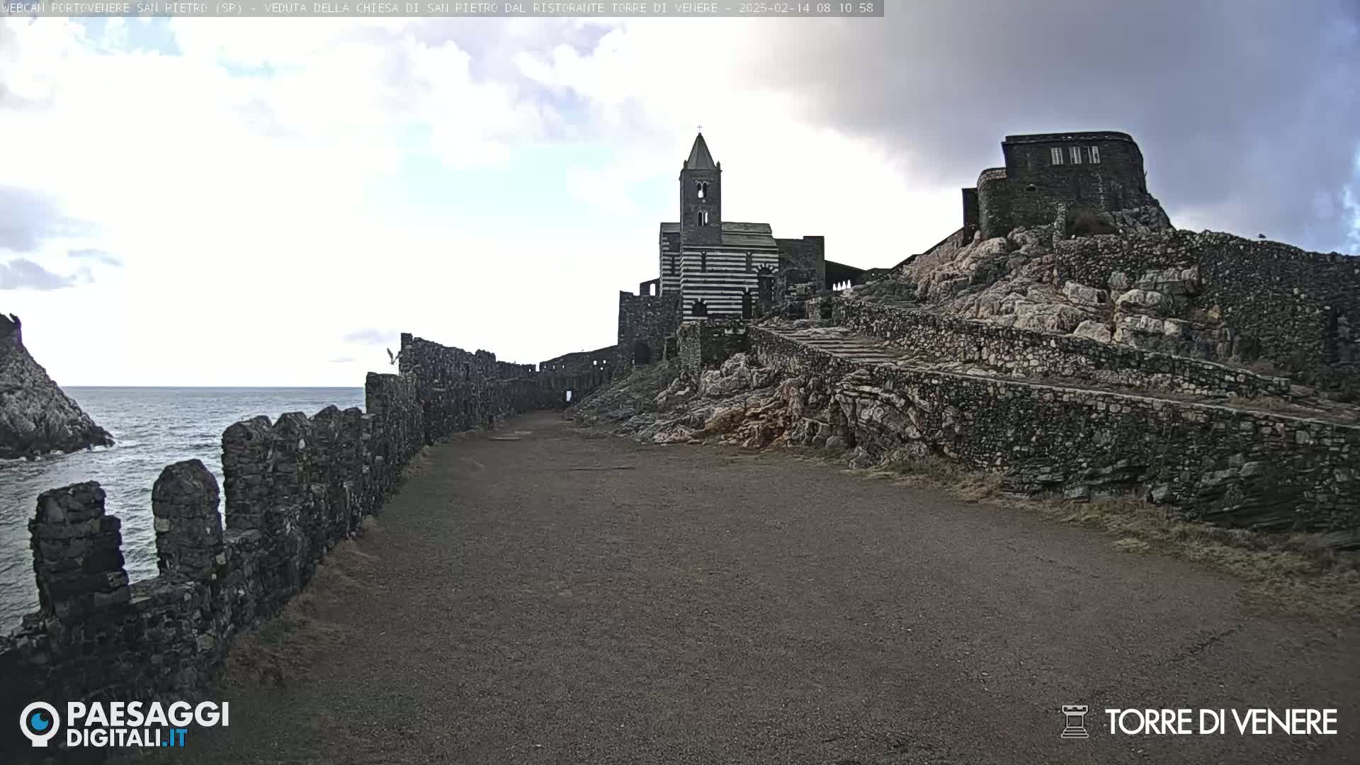 A stone path leads to a striped stone church situated on a rocky hill overlooking a calm sea under a cloudy sky.