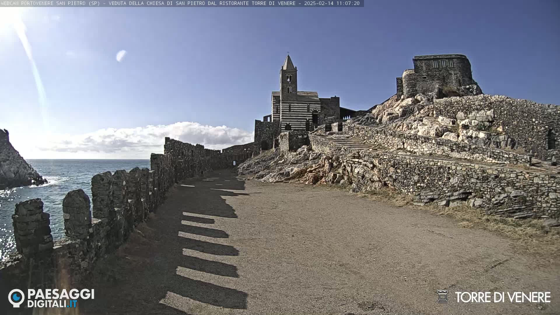 A stone pathway leads to a striped church and stone ruins perched on a rocky cliff overlooking a calm sea under a mostly clear, sunny sky.