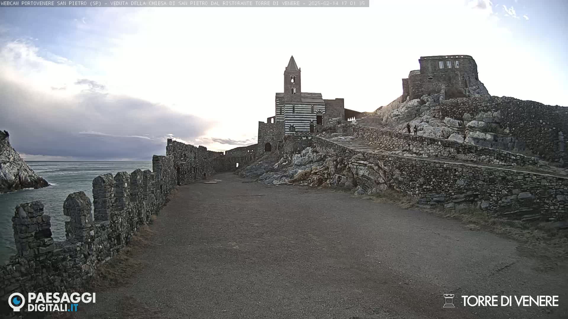 A stone path leads to a striped church and castle ruins perched on a rocky cliff overlooking a calm sea under a partly cloudy sky.