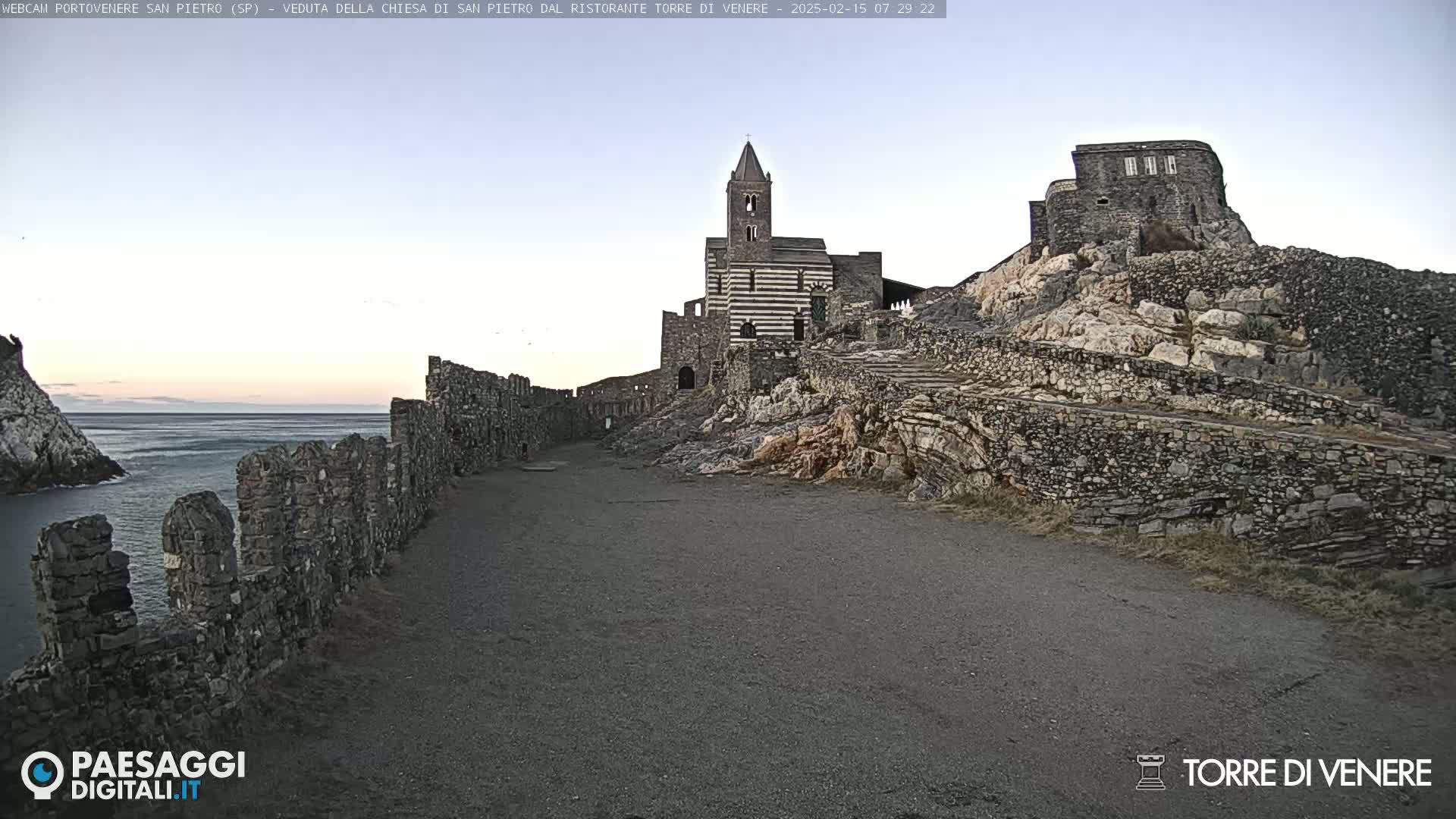 A stone path leads toward a striped church and castle ruins perched atop a rocky hill overlooking a calm sea under a clear, pale sky.
