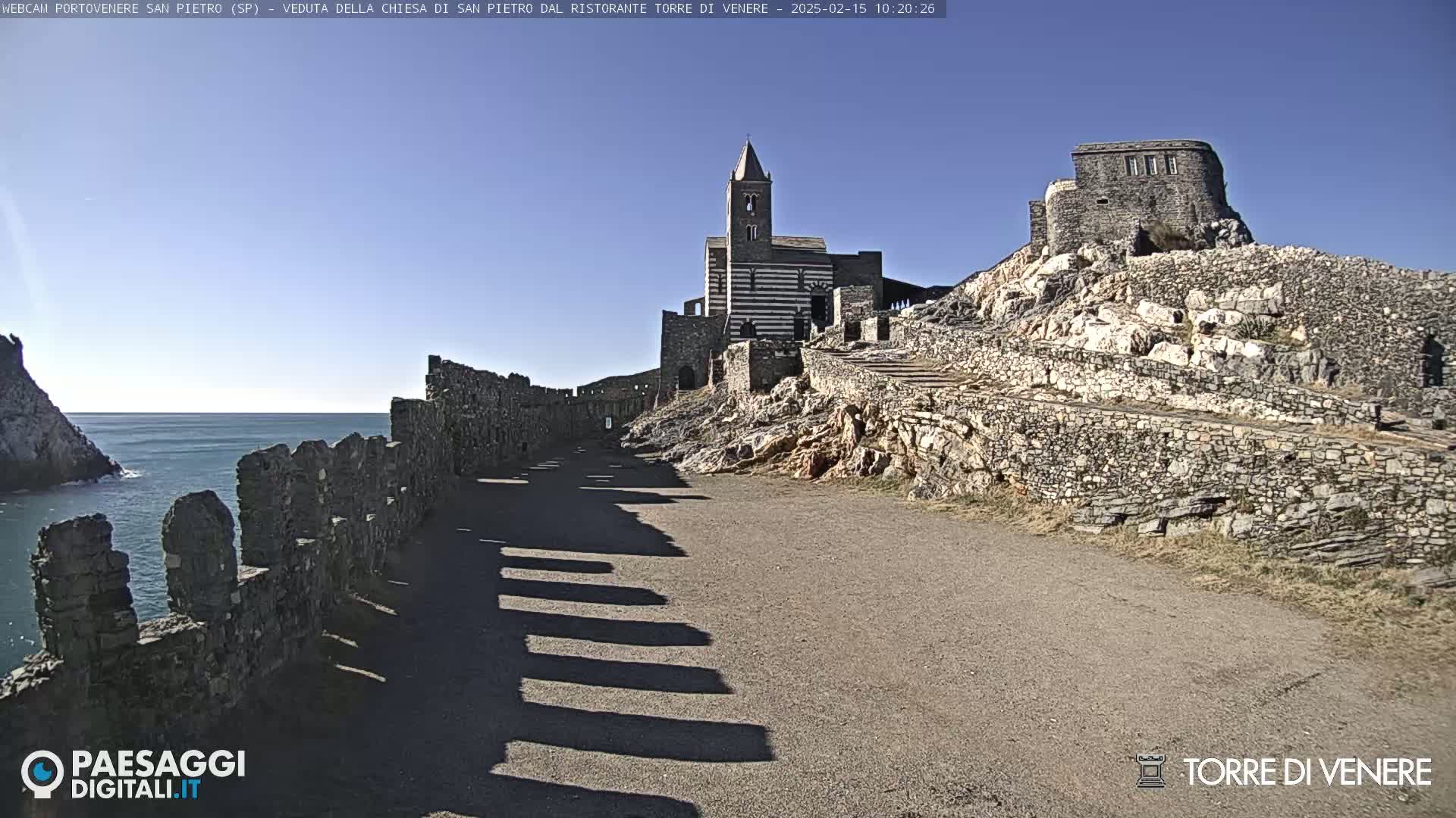 A stone pathway leads toward a striped church and castle ruins overlooking a calm ocean under a clear blue sky.
