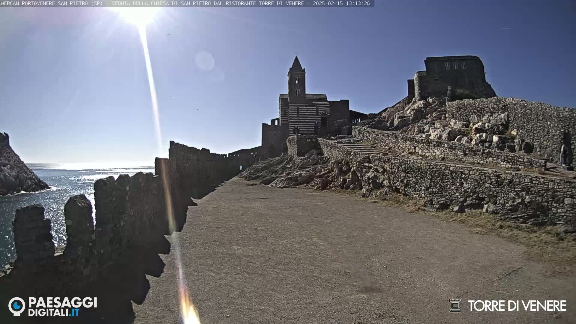 A sunny day reveals a stone path leading to a striped church and ruins situated on a rocky hillside overlooking the ocean.