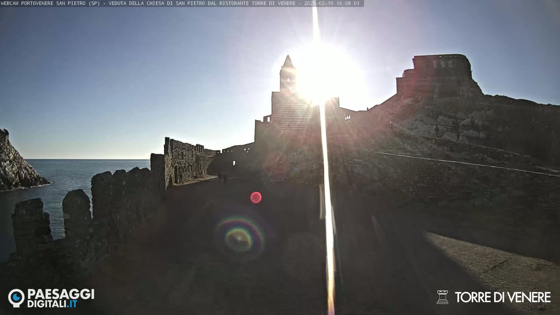 A sunlit view of a stone church and castle ruins on a rocky coast overlooking the sea on a bright, clear day.