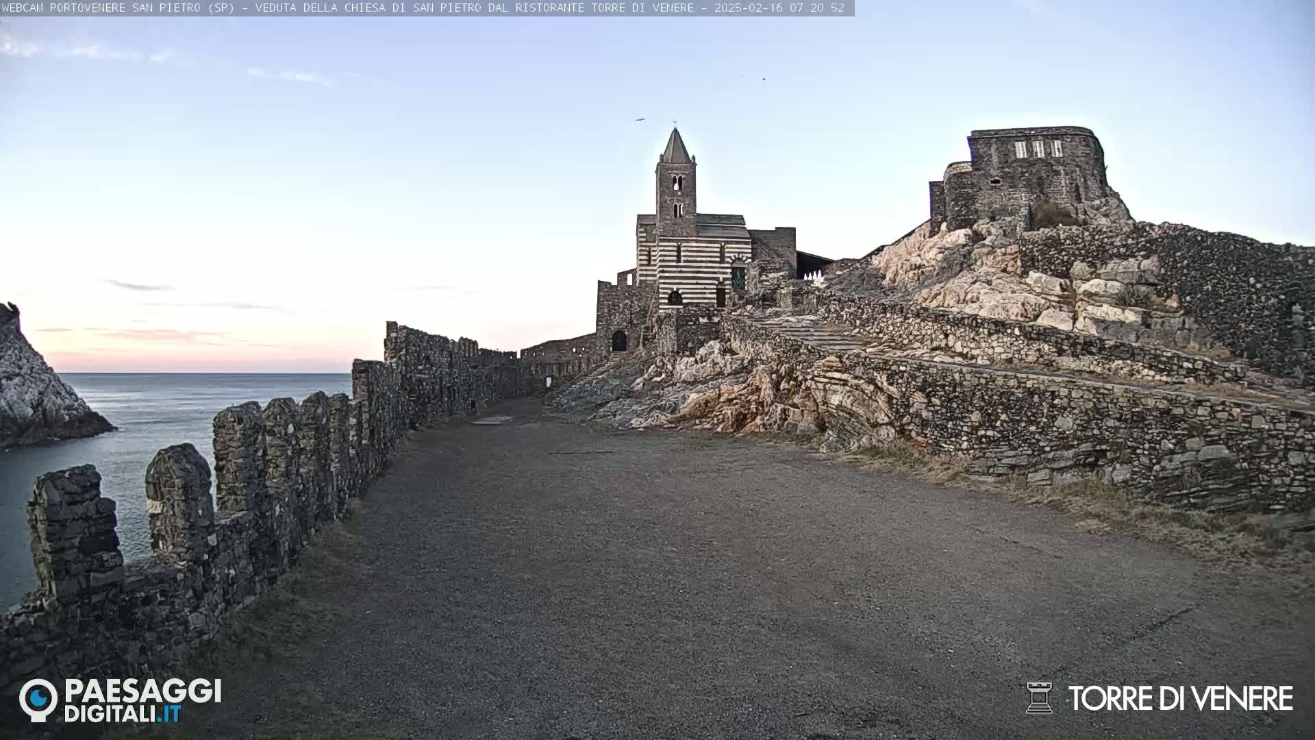 A stone church and castle ruins on a rocky hill overlook a calm sea under a clear, pale sky.