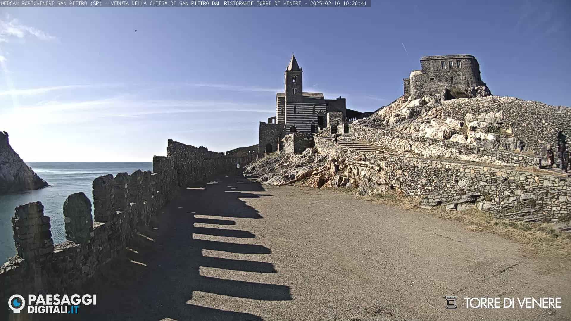 A stone pathway leads to a striped church and castle ruins perched on a rocky cliff overlooking a calm sea under a clear sky.