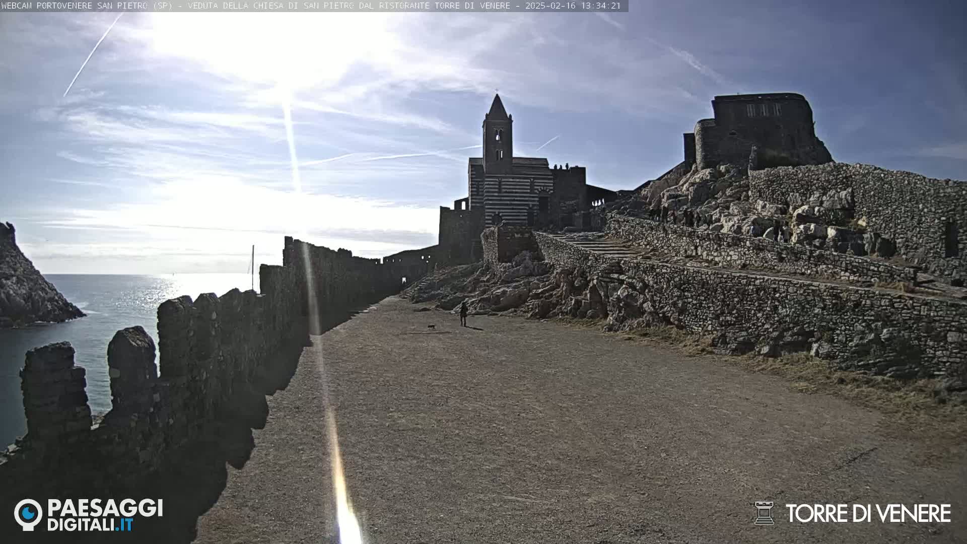 A sunny day reveals a coastal stone pathway leading to a church and castle ruins, with the ocean visible in the background.