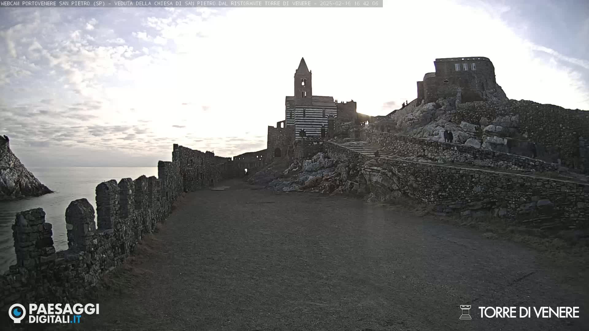 Stone ruins and a striped church stand on a rocky hillside overlooking a calm sea under a partly cloudy sky.
