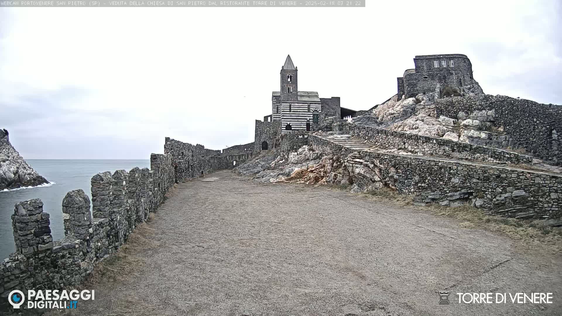 A stone path leads toward a church and stone structures situated on a rocky hill overlooking a calm sea under an overcast sky.