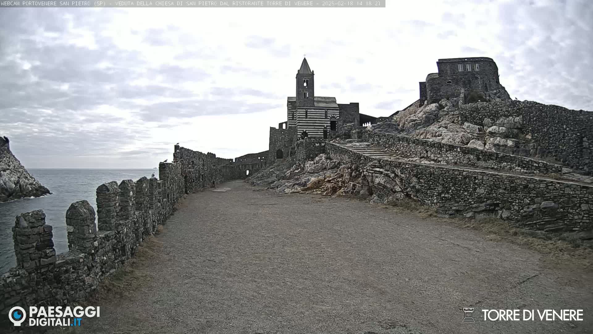A stone path leads to a striped church and castle ruins on a rocky hill overlooking a calm ocean under a cloudy sky.