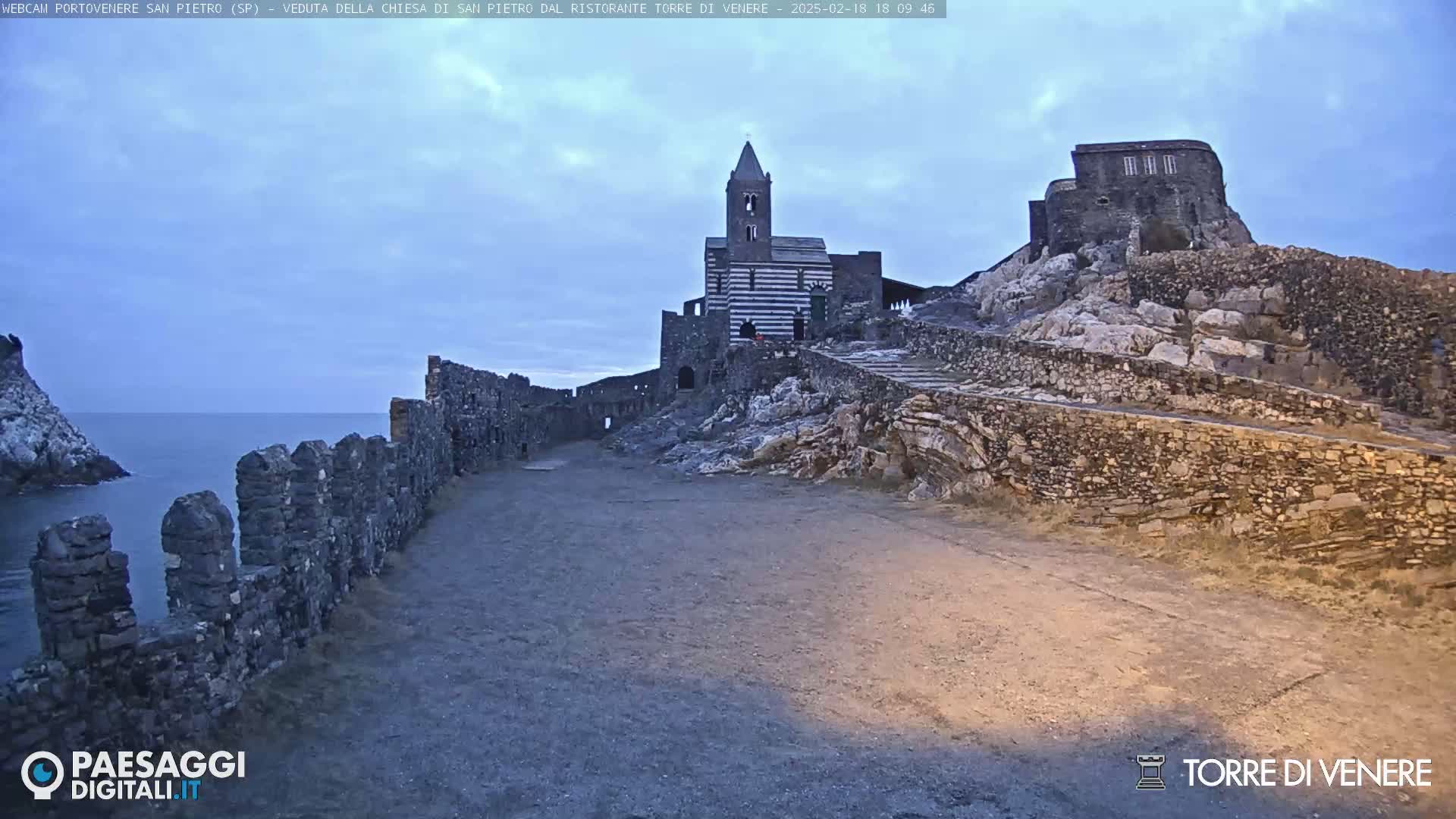 A stone church and castle ruins sit atop a rocky cliff overlooking a calm ocean under a partly cloudy twilight sky.