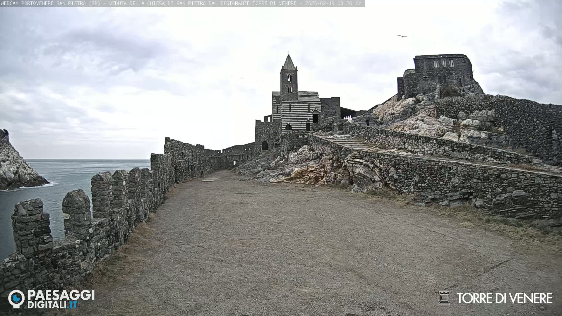 A stone church and castle ruins sit atop a rocky cliff overlooking a calm ocean under a cloudy sky.