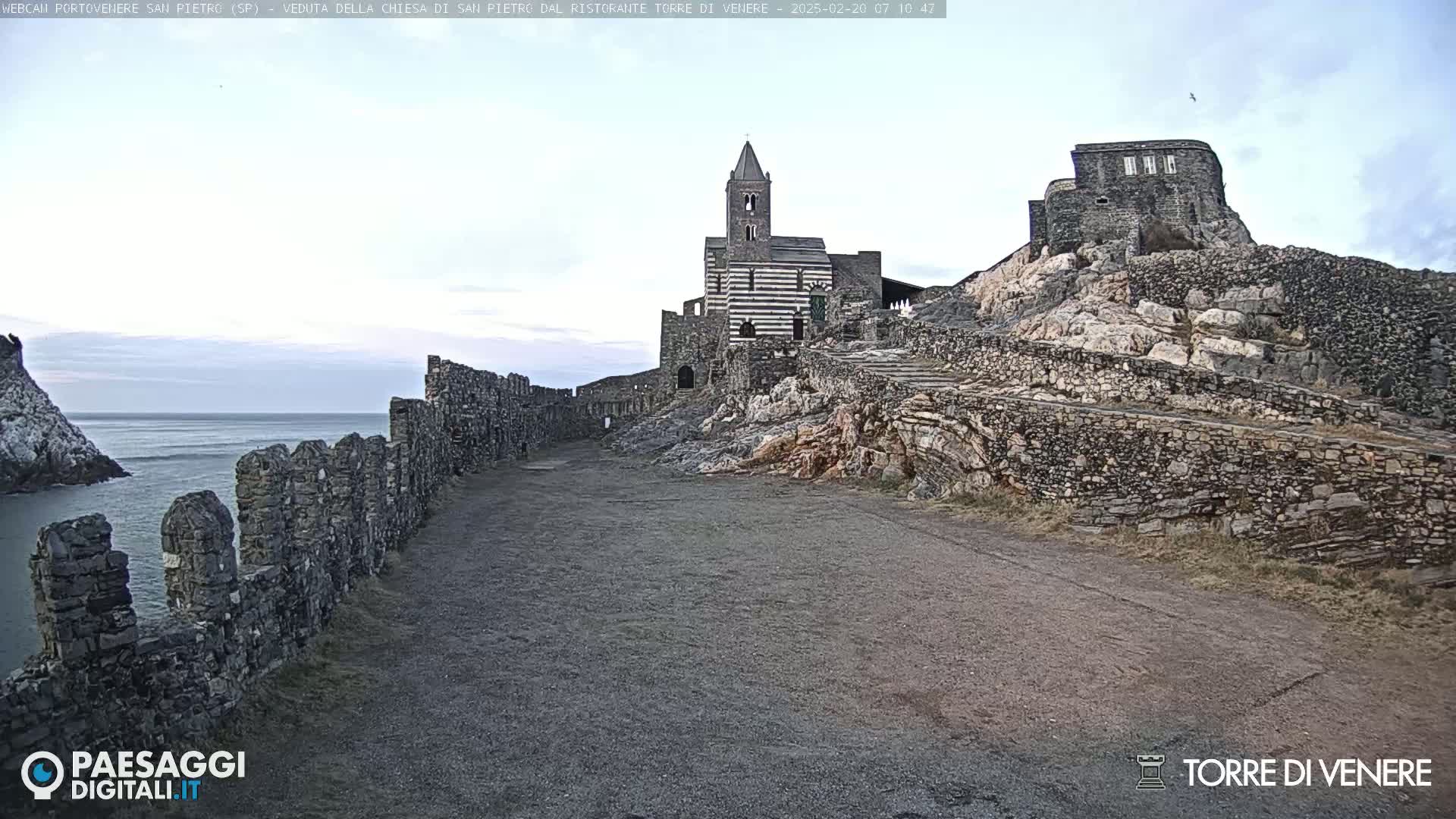 A stone pathway leads to a church and castle ruins perched atop a rocky cliff overlooking a calm sea under a mostly clear sky.