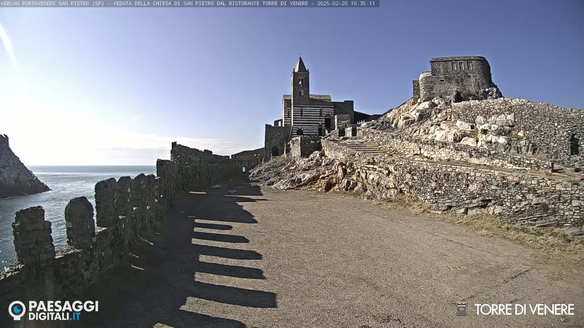 A stone pathway leads to a striped church and castle ruins perched on a rocky cliff overlooking a calm sea under a clear blue sky.