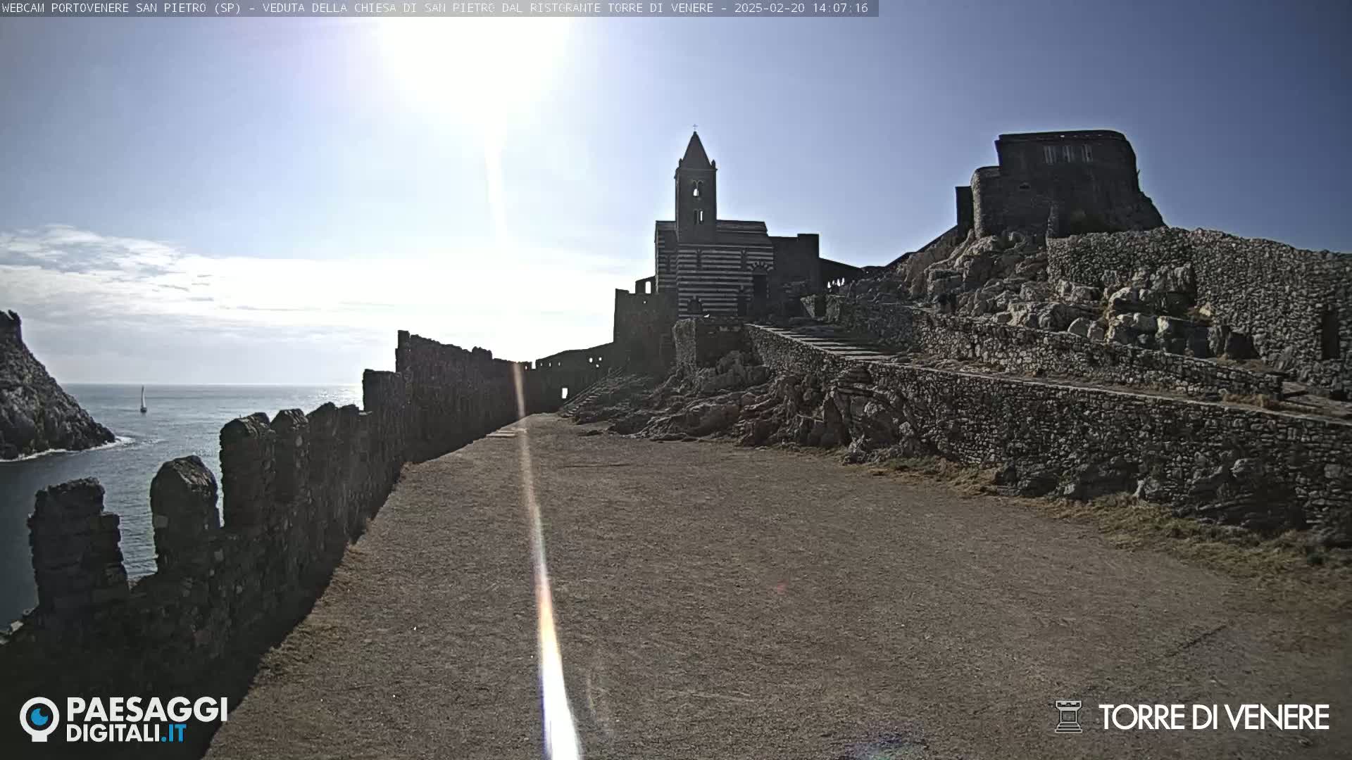 A stone pathway leads to a striped church and castle ruins overlooking a calm ocean under a mostly sunny sky.