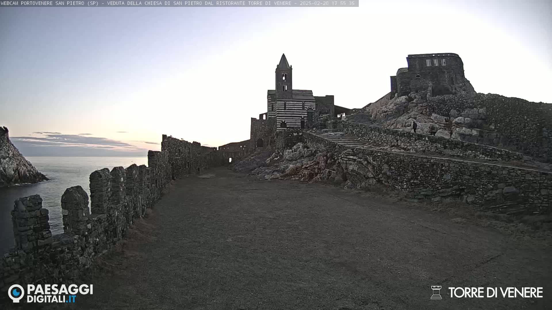 A stone path leads to a striped church and castle ruins perched on a rocky cliff overlooking a calm sea at dawn.