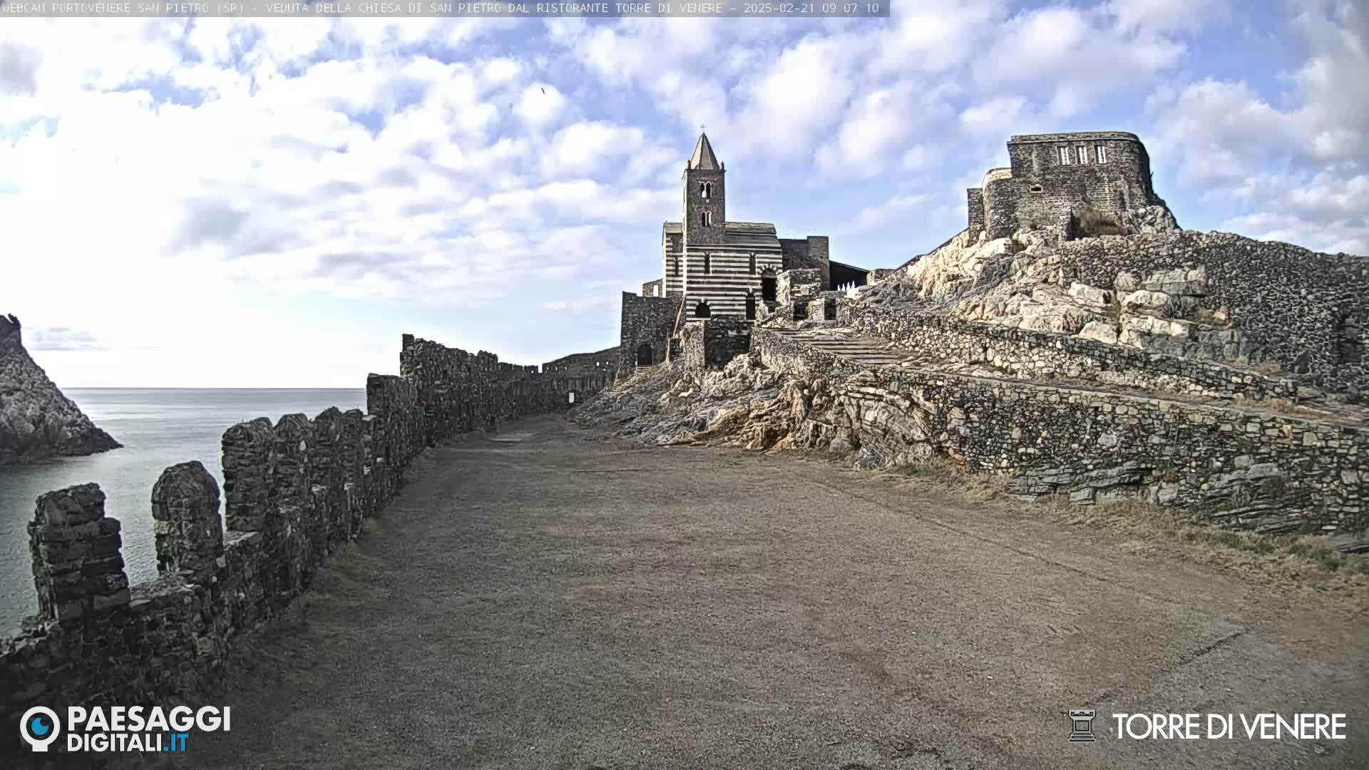 A partly cloudy day reveals a stone church and ruins overlooking a calm sea, with ancient stone walls extending along the foreground.