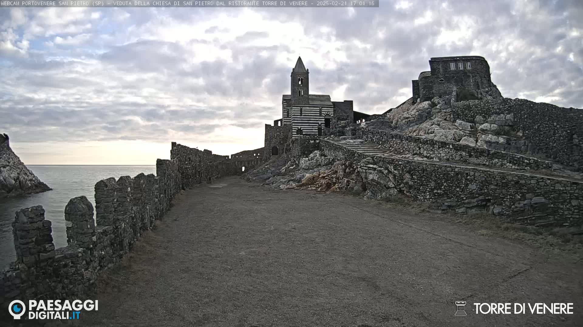Stone ruins and a striped church stand on a rocky hill overlooking a calm sea under a cloudy sky.