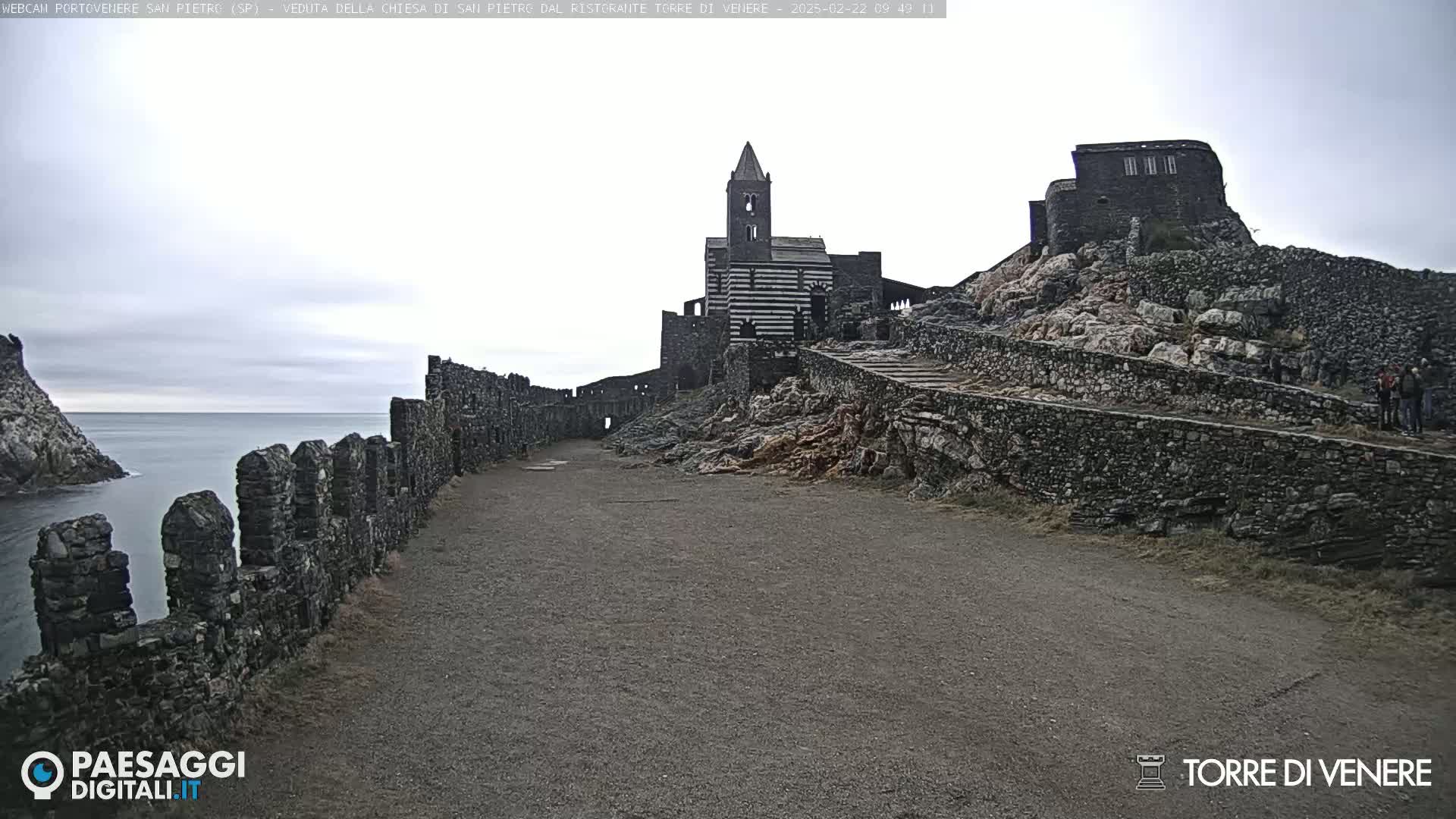 A stone path leads toward a church and castle ruins situated on a rocky hillside overlooking a calm sea under an overcast sky.