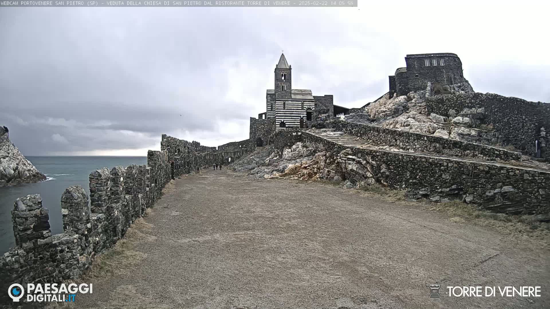 A stone path leads toward a striped church and stone buildings atop a rocky hill overlooking a calm sea under a cloudy sky.