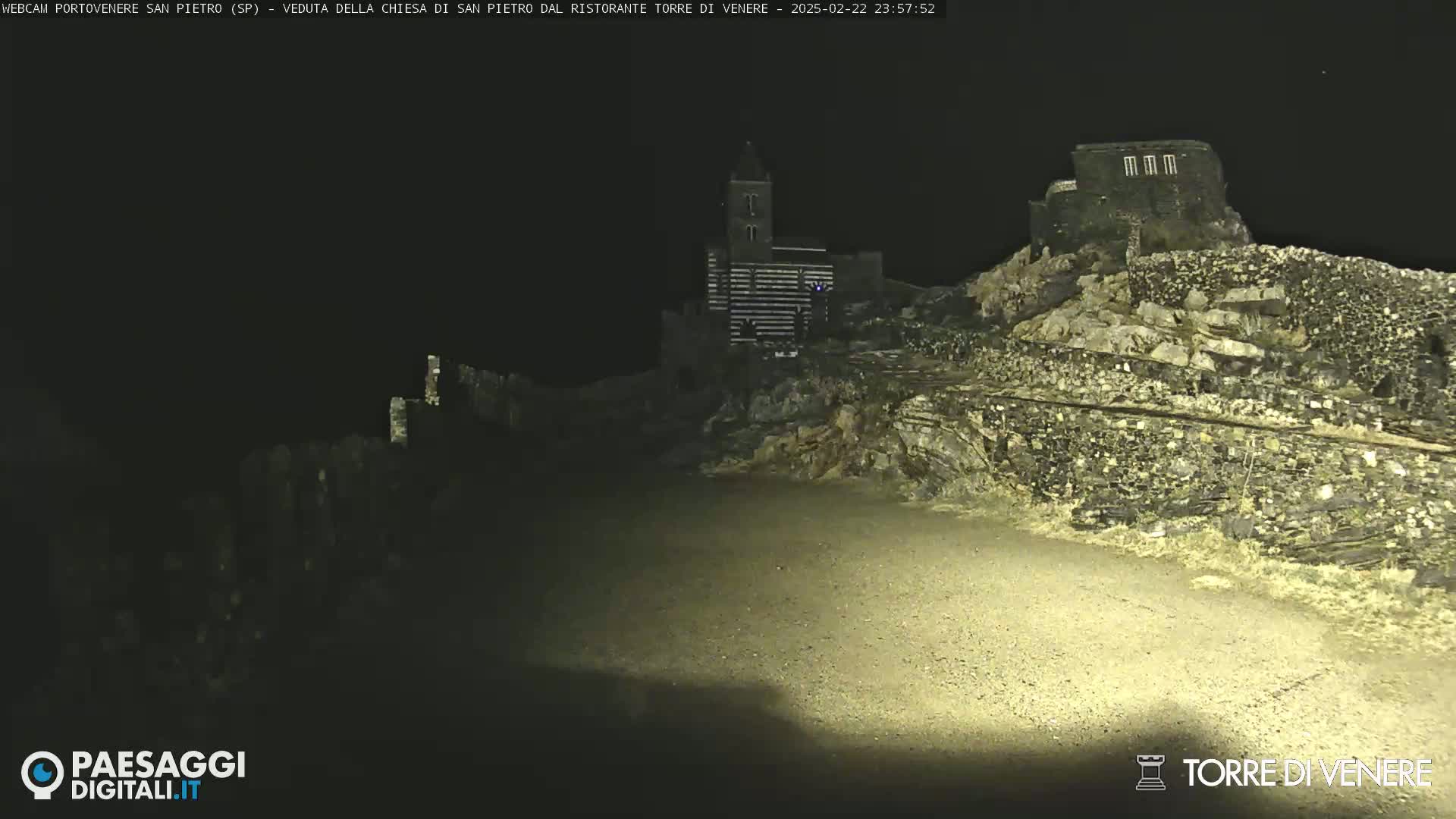 At night, stone structures, including a church and what appears to be a wall or fortification, are illuminated on a hilltop.