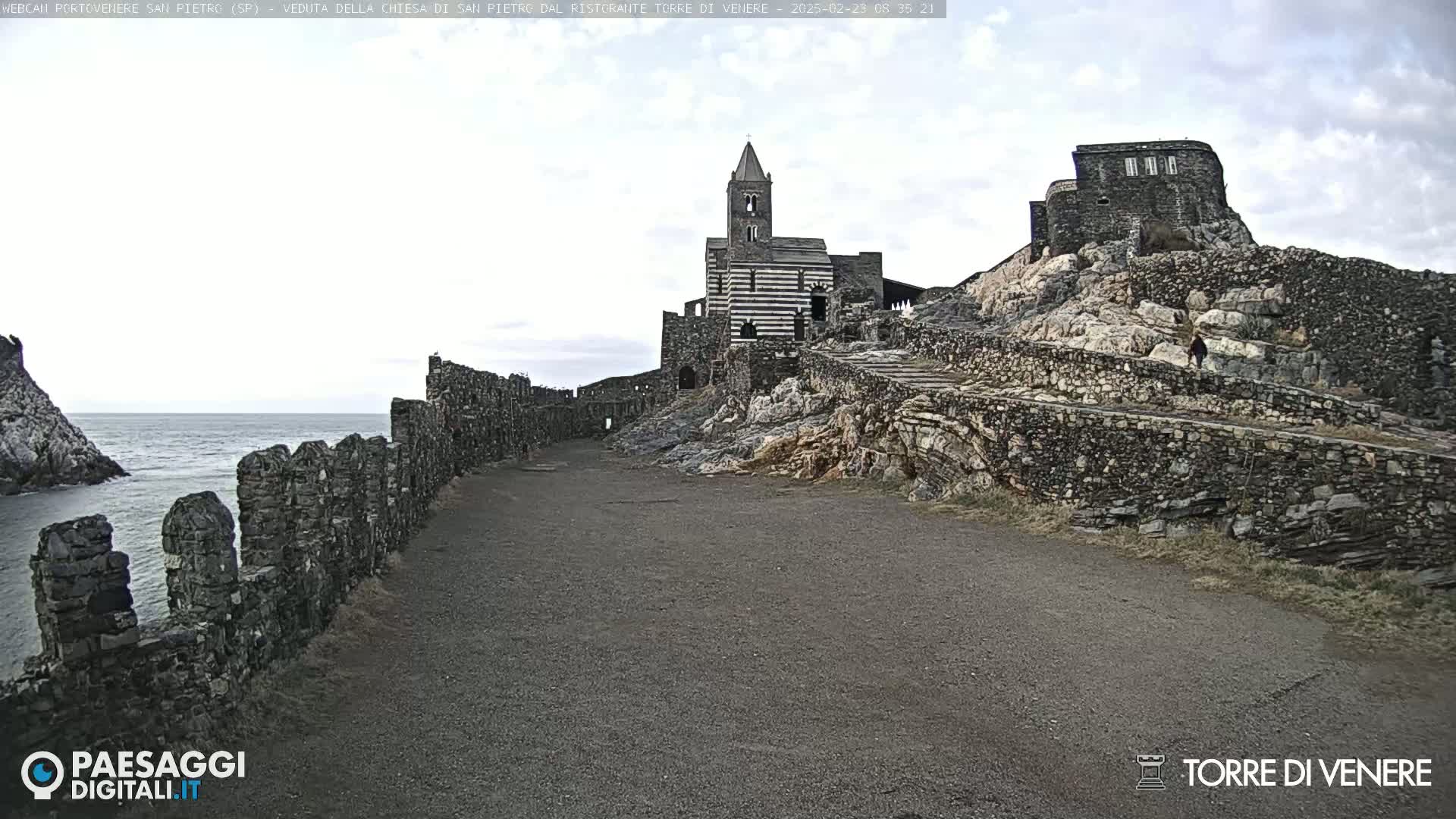 A stone church and castle ruins sit atop a rocky hill overlooking a calm ocean under a cloudy sky.