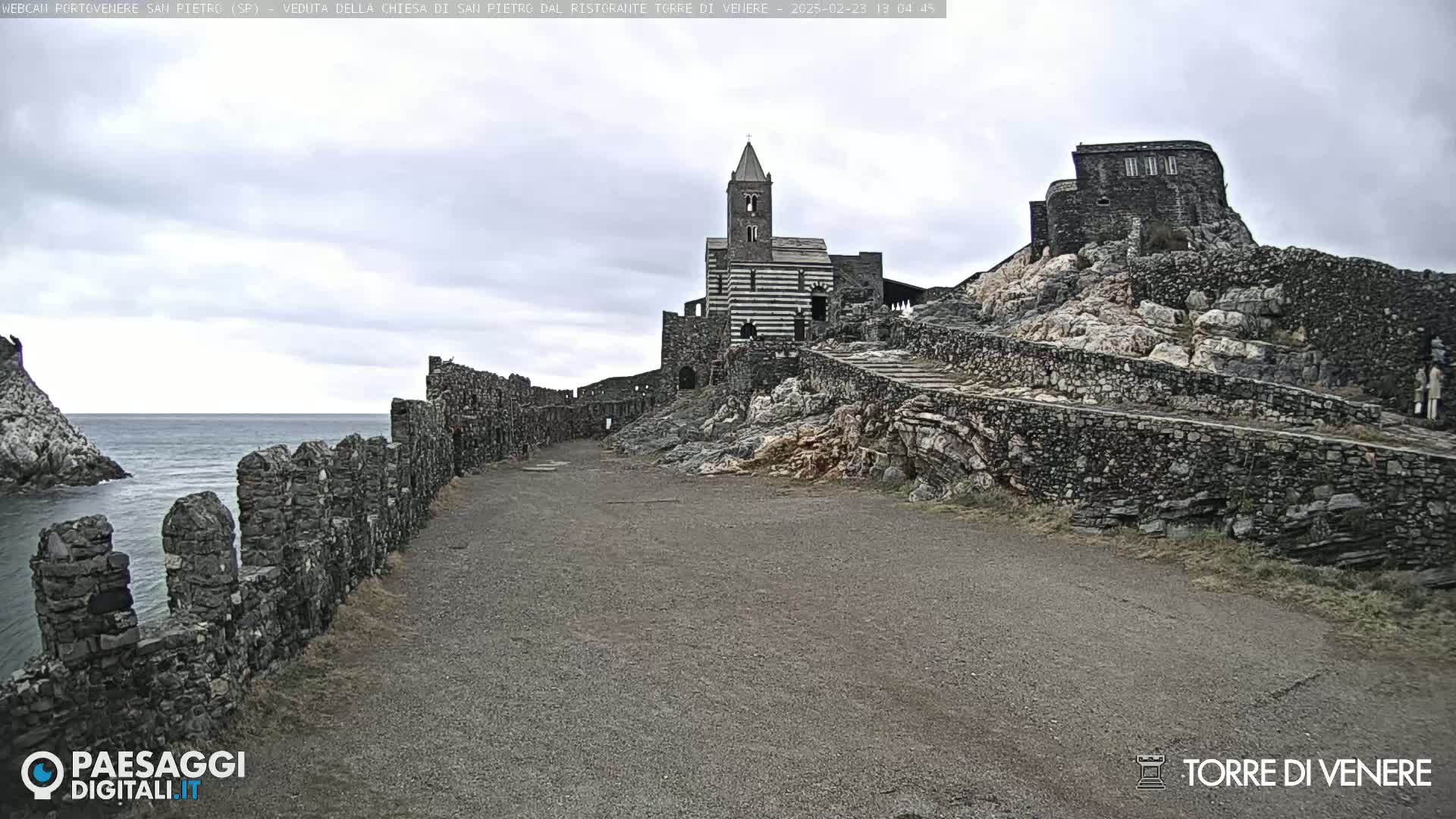 A stone path leads to a church and castle ruins perched on a rocky outcrop overlooking a calm sea under an overcast sky.