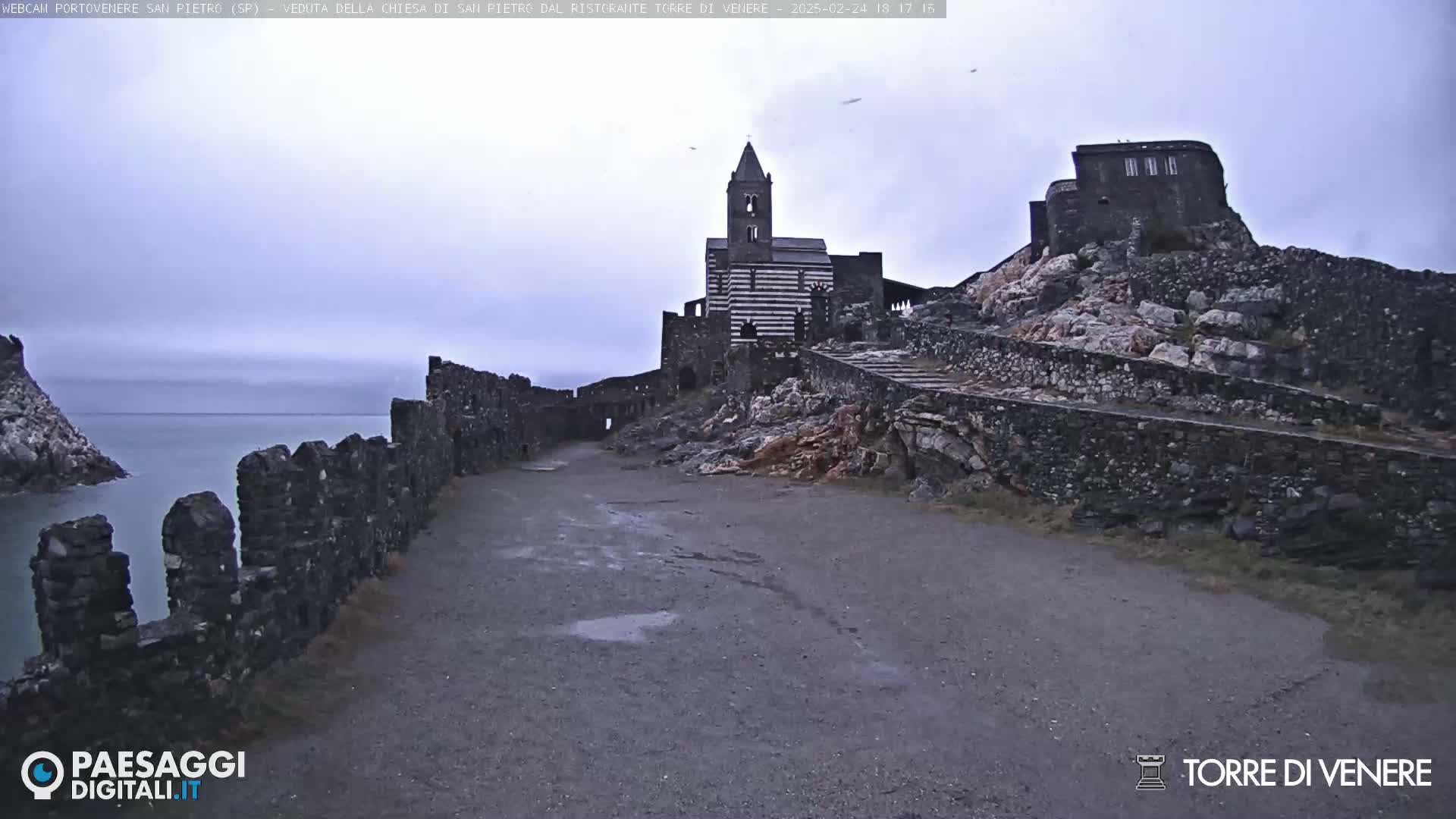 A stone path leads to a striped church and stone buildings atop a rocky cliff overlooking a calm sea under an overcast sky.