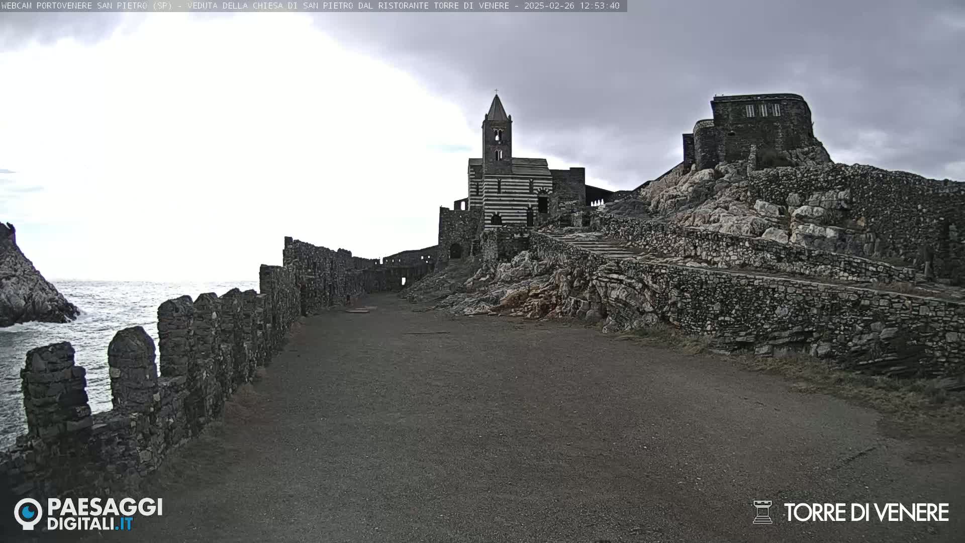 A stone church and ruins sit atop a rocky cliff overlooking a dark, choppy sea under an overcast sky.