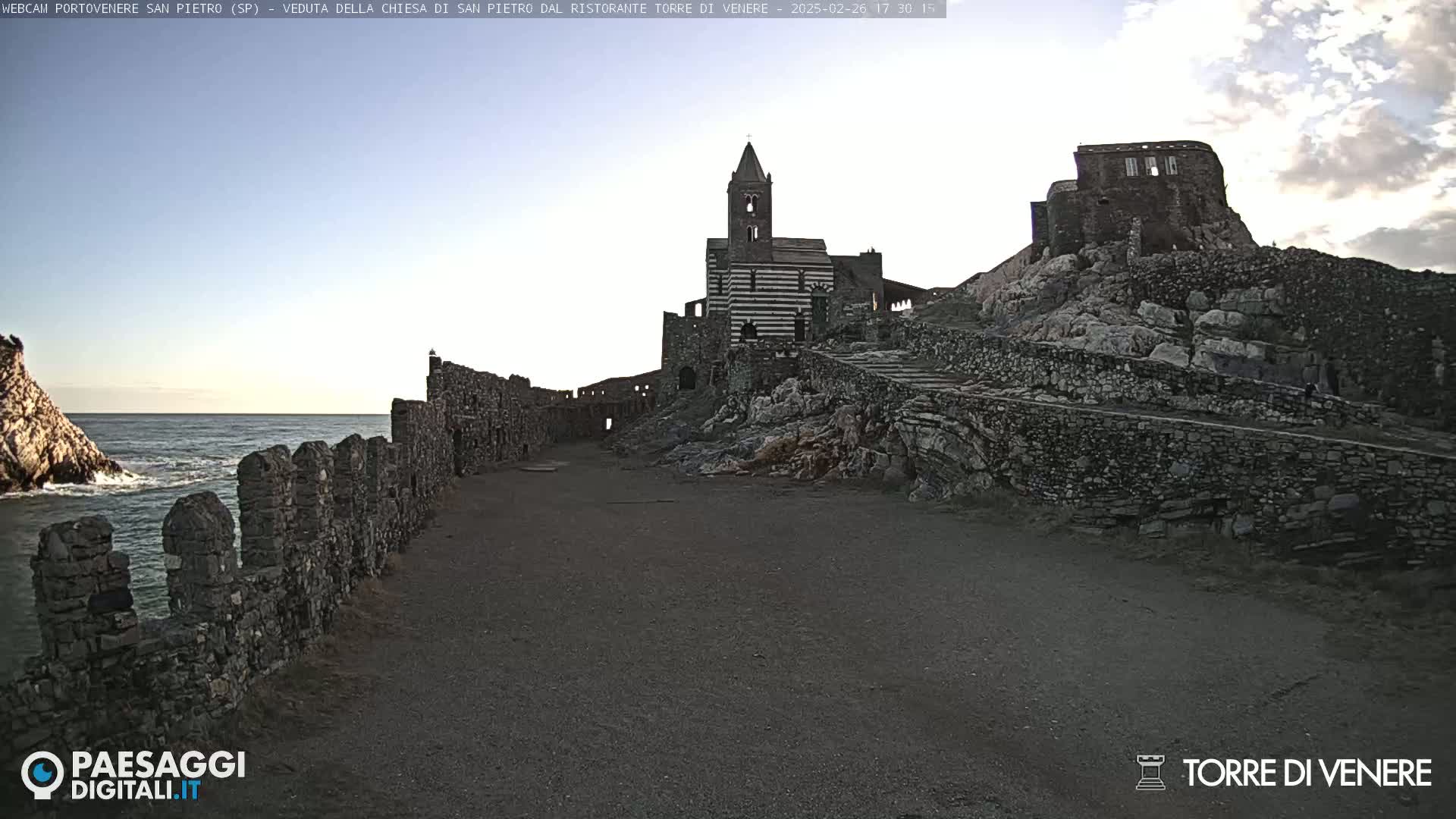 A stone path leads to a striped church and ruins on a rocky hill overlooking a calm sea under a clear sky.