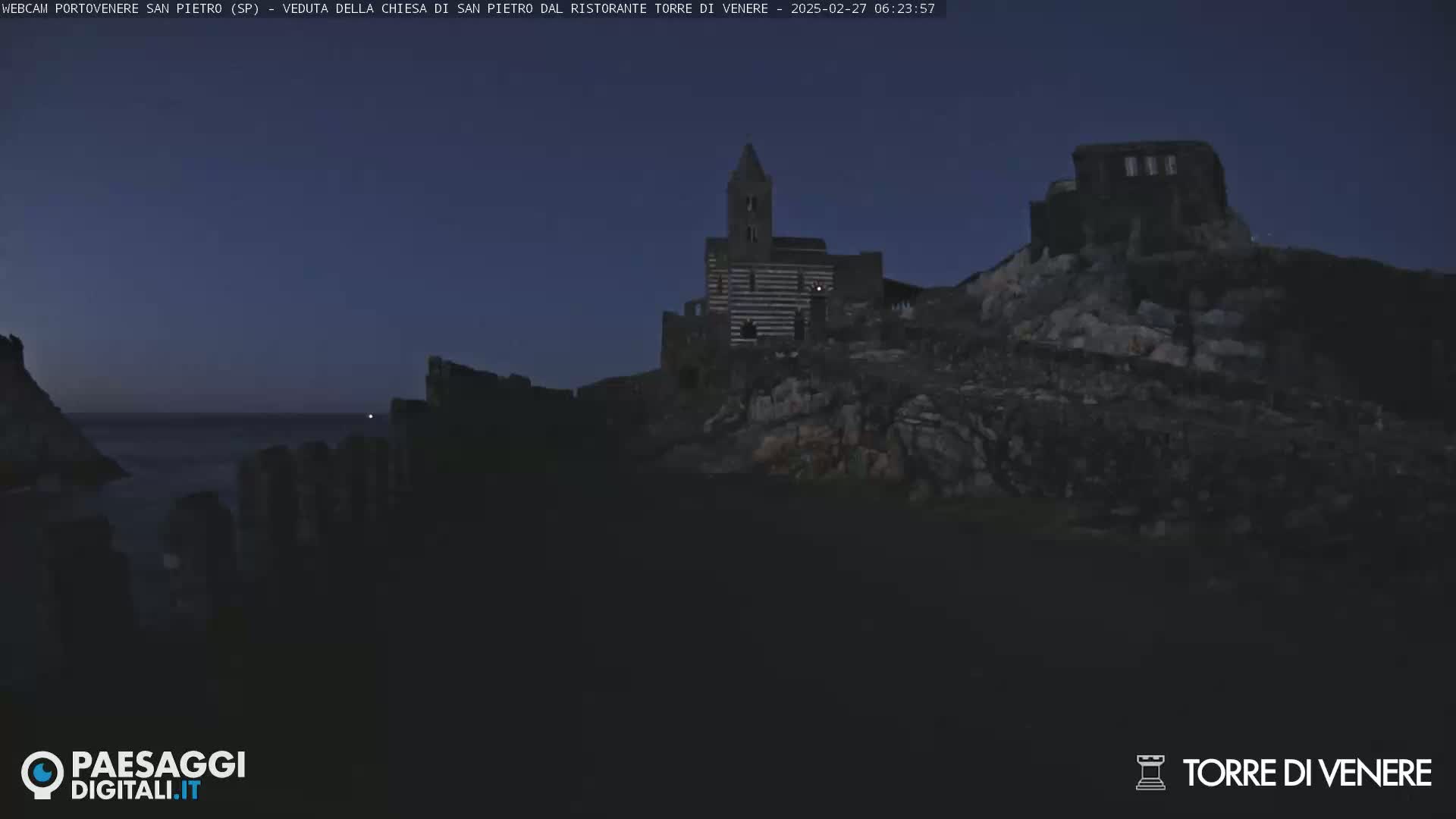 A dark, rocky coastal area at twilight features a striped stone church and other buildings on a hilltop overlooking the sea.
