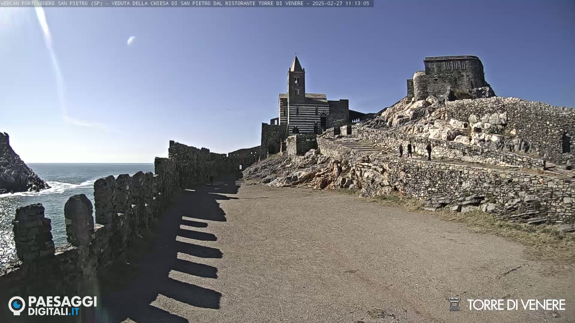 A stone path leads toward a striped church and castle ruins perched atop a rocky cliff overlooking a calm ocean under a clear, sunny sky.
