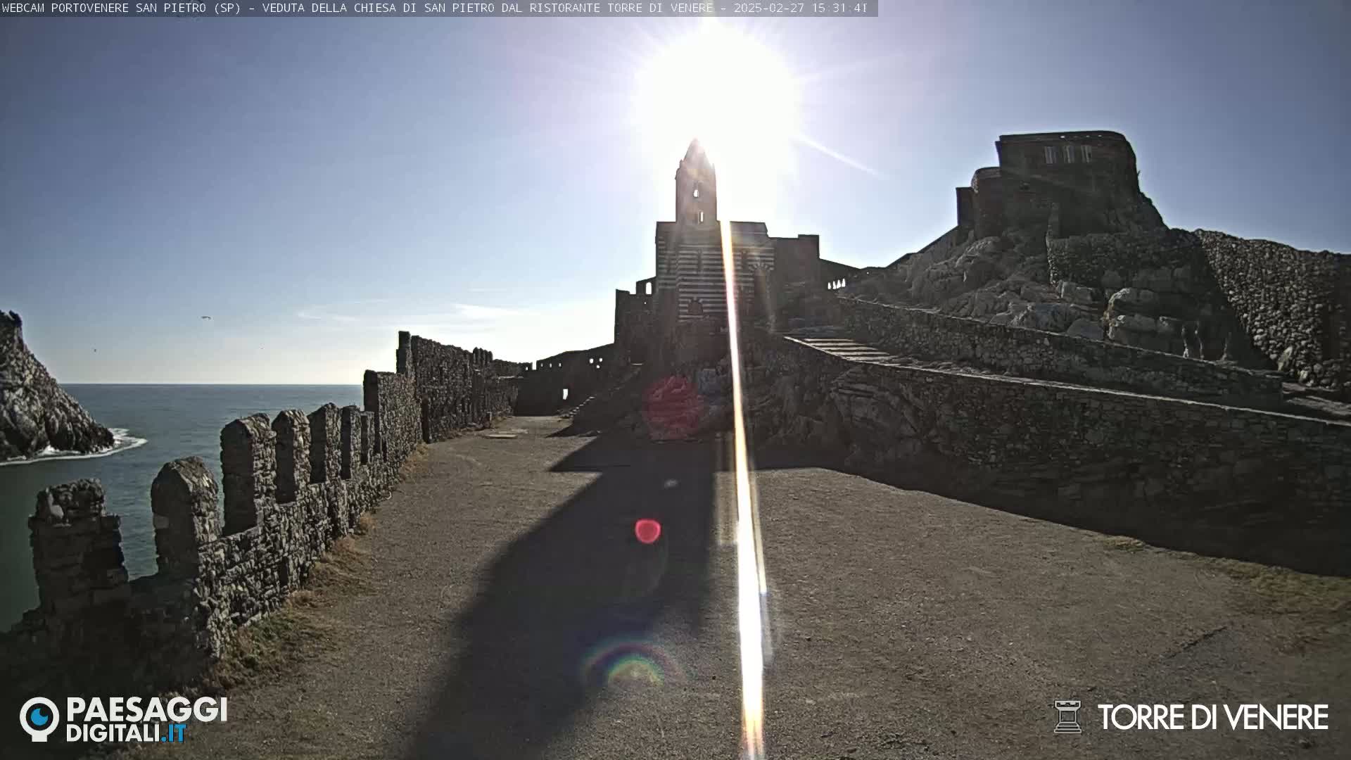 A stone church and buildings sit atop a rocky cliff overlooking a calm sea under a bright, sunny sky.