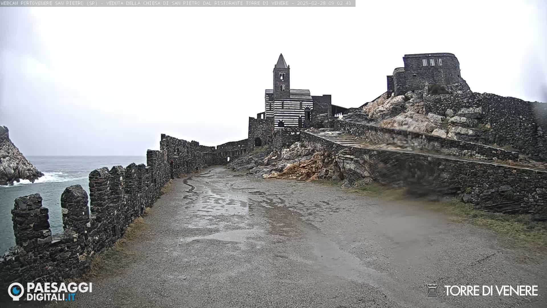A stone pathway leads to a striped stone church and adjacent stone structures on a rocky outcrop overlooking a gray, overcast ocean.