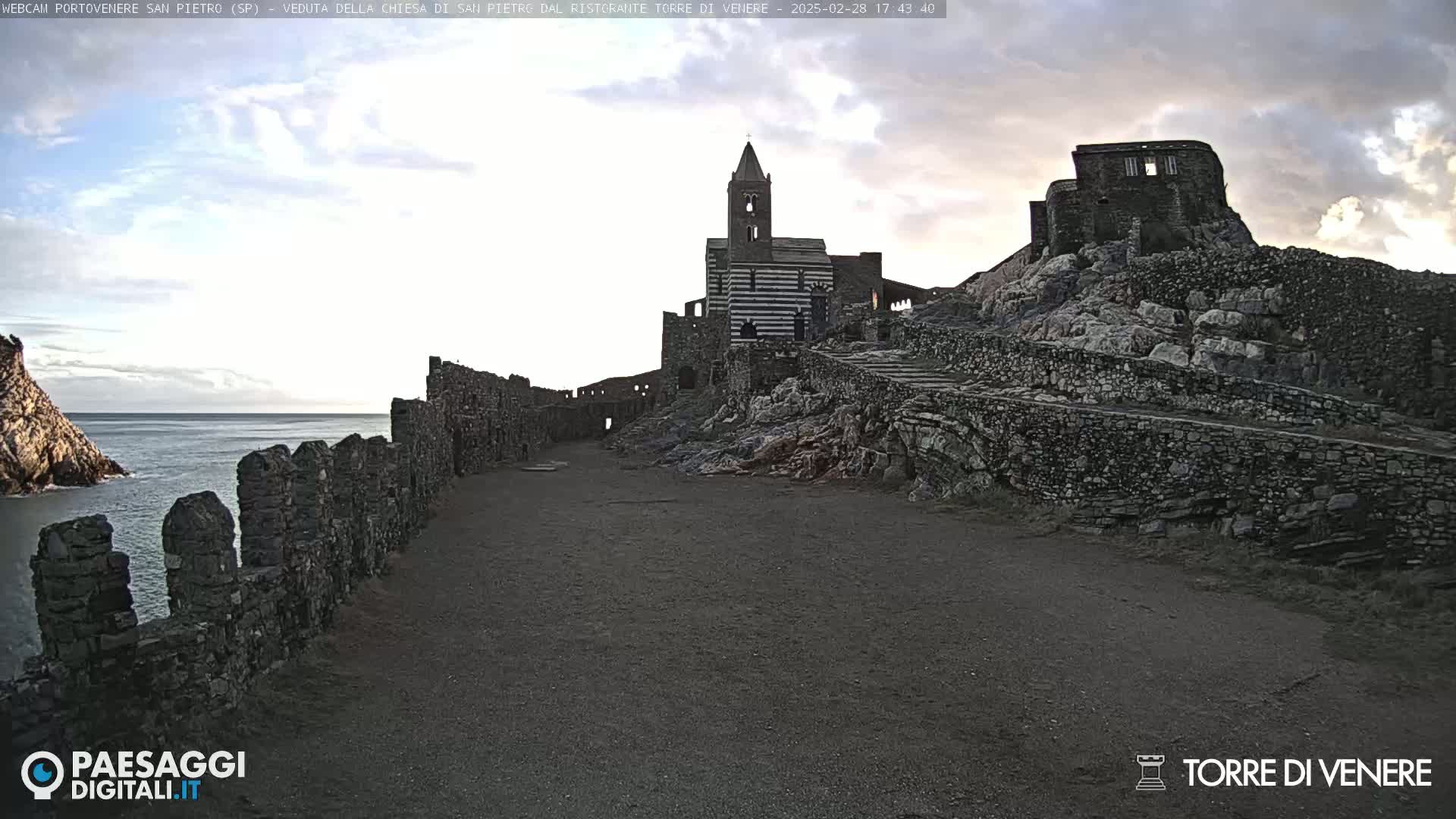 A stone pathway leads to a striped church and ruined buildings on a rocky hill overlooking a calm sea under a partly cloudy sky.