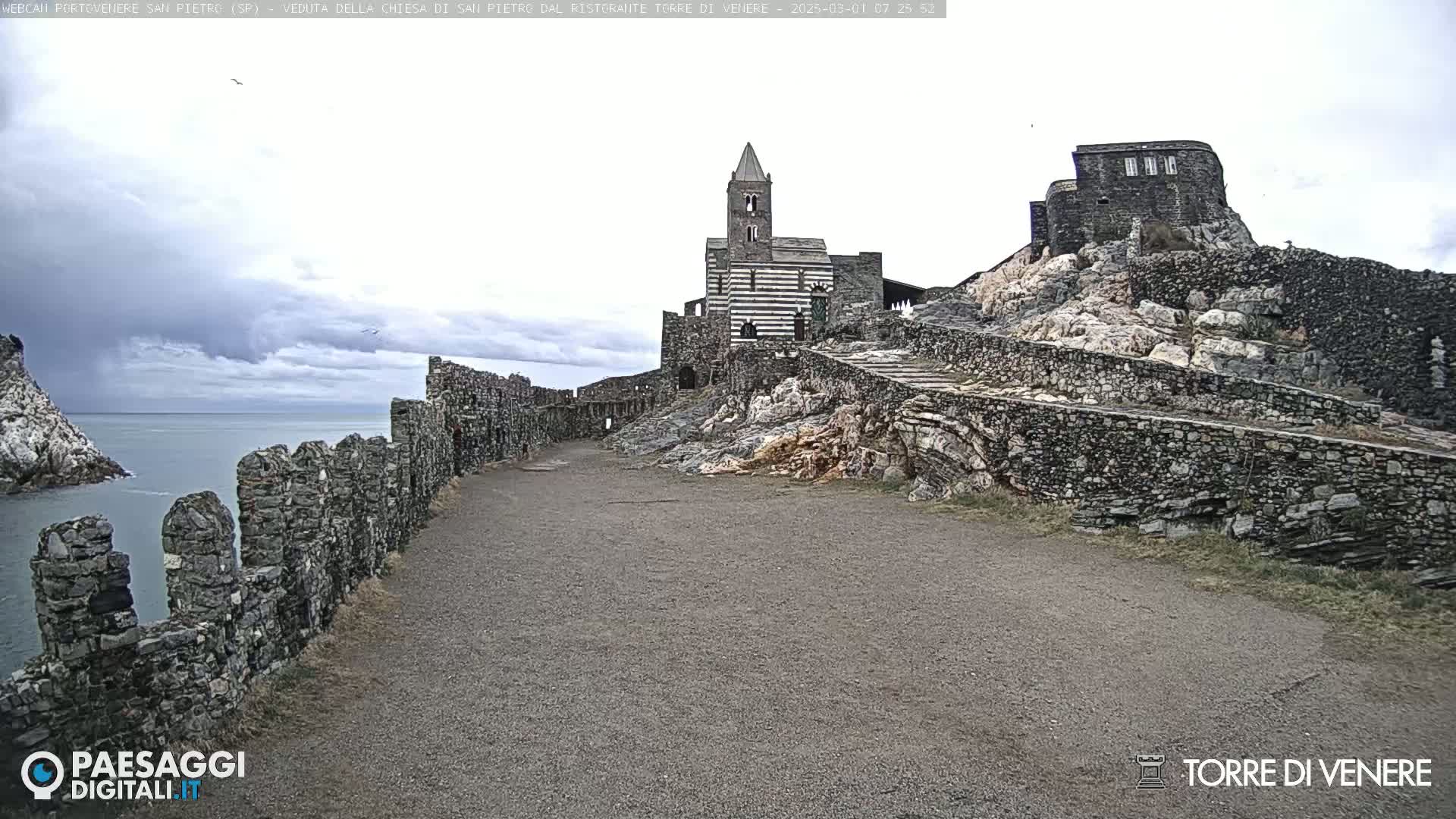 A stone pathway leads to a striped stone church atop a rocky cliff overlooking a calm sea under a cloudy sky.
