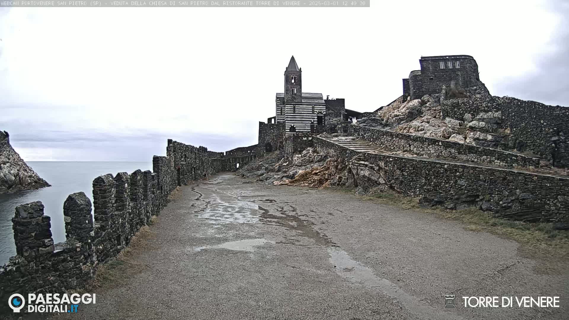 A stone pathway leads to a striped stone church and fortress on a rocky cliff overlooking a calm sea under an overcast sky.