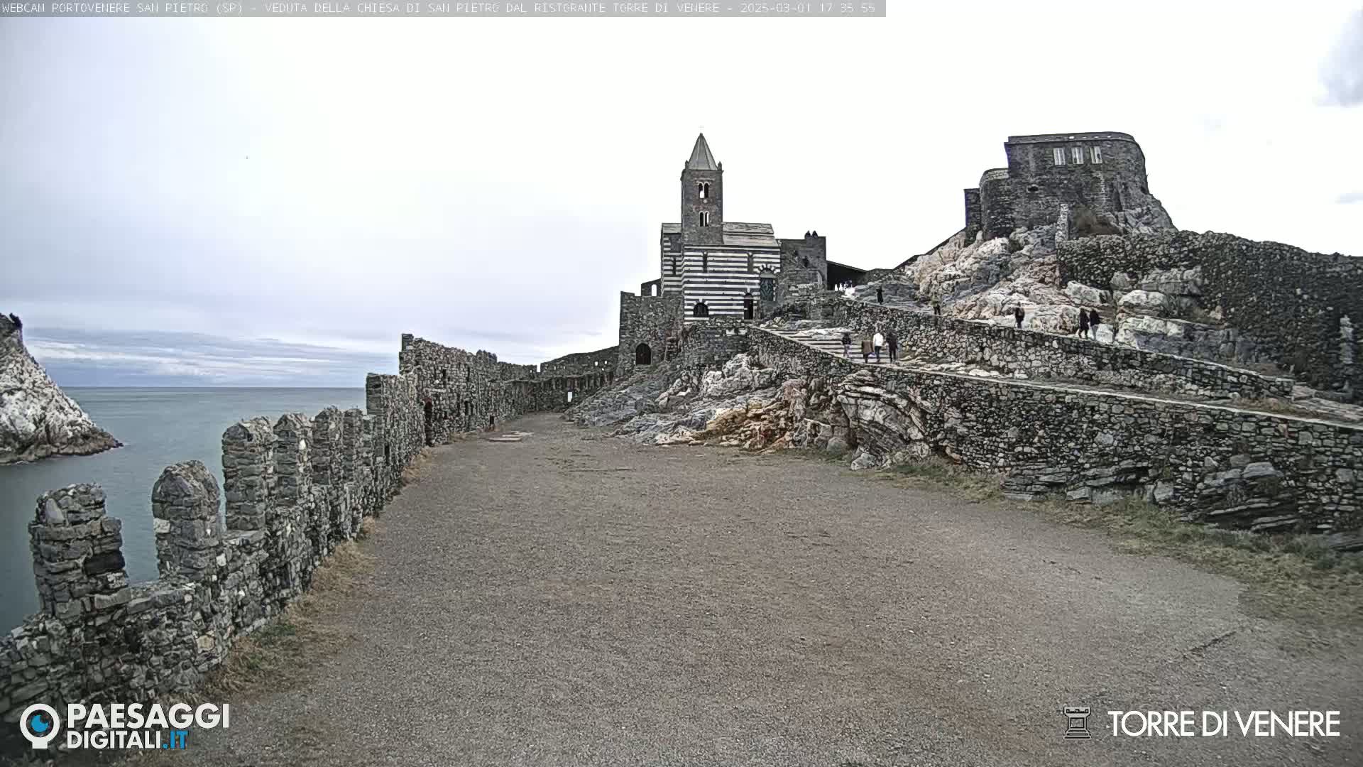 A stone pathway leads to a striped stone church and castle ruins on a rocky cliff overlooking a calm ocean under an overcast sky.
