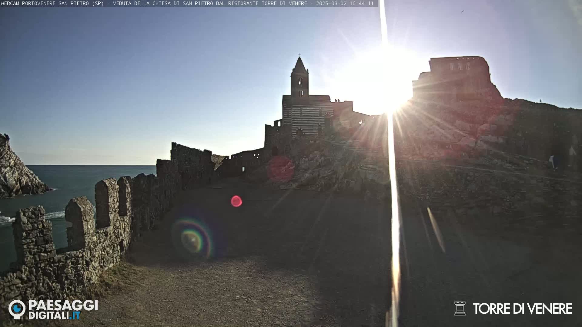A rocky coastal area with a church and stone walls is bathed in bright sunlight, overlooking a calm sea.