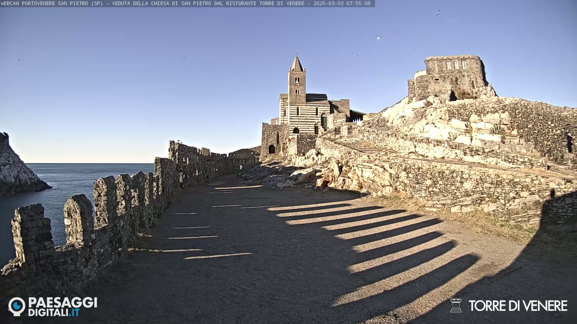 A stone pathway leads to a striped church and stone ruins on a sunny day overlooking a calm ocean.