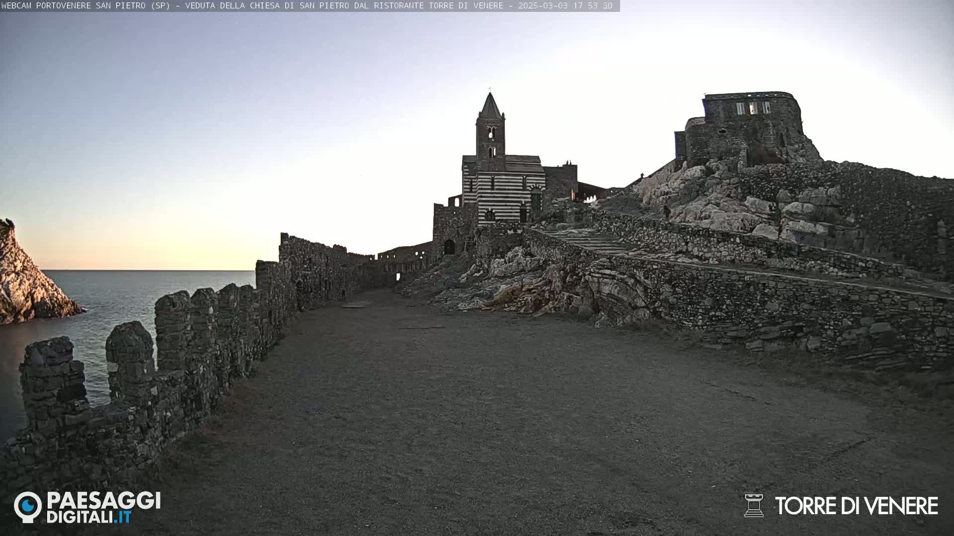 A coastal stone path leads to a striped church and ruins atop a rocky cliff overlooking a calm sea under a twilight sky.