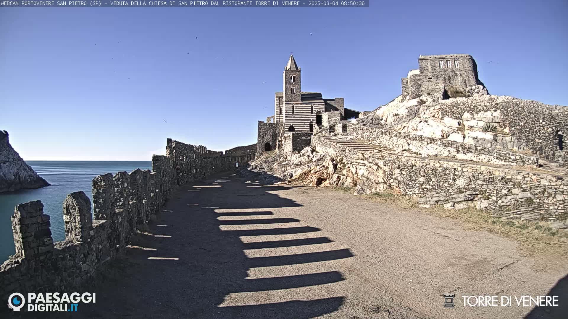 A stone church and ruins sit atop a rocky cliff overlooking a calm ocean under a clear blue sky.