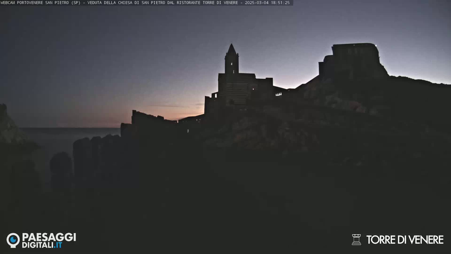 A dark silhouette of a church and castle-like structures sits atop a hill overlooking a calm sea at twilight.