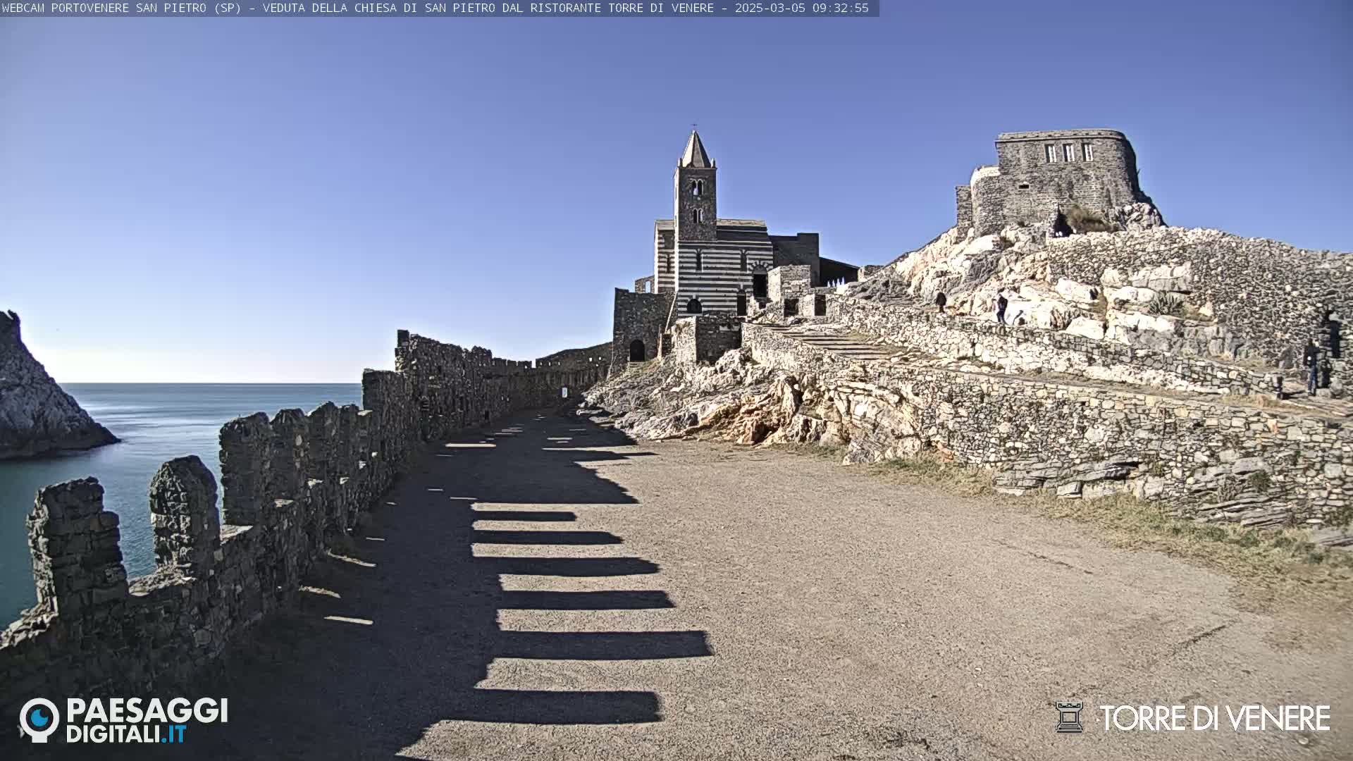 A stone pathway leads to a striped church and castle ruins overlooking a calm sea under a clear blue sky.