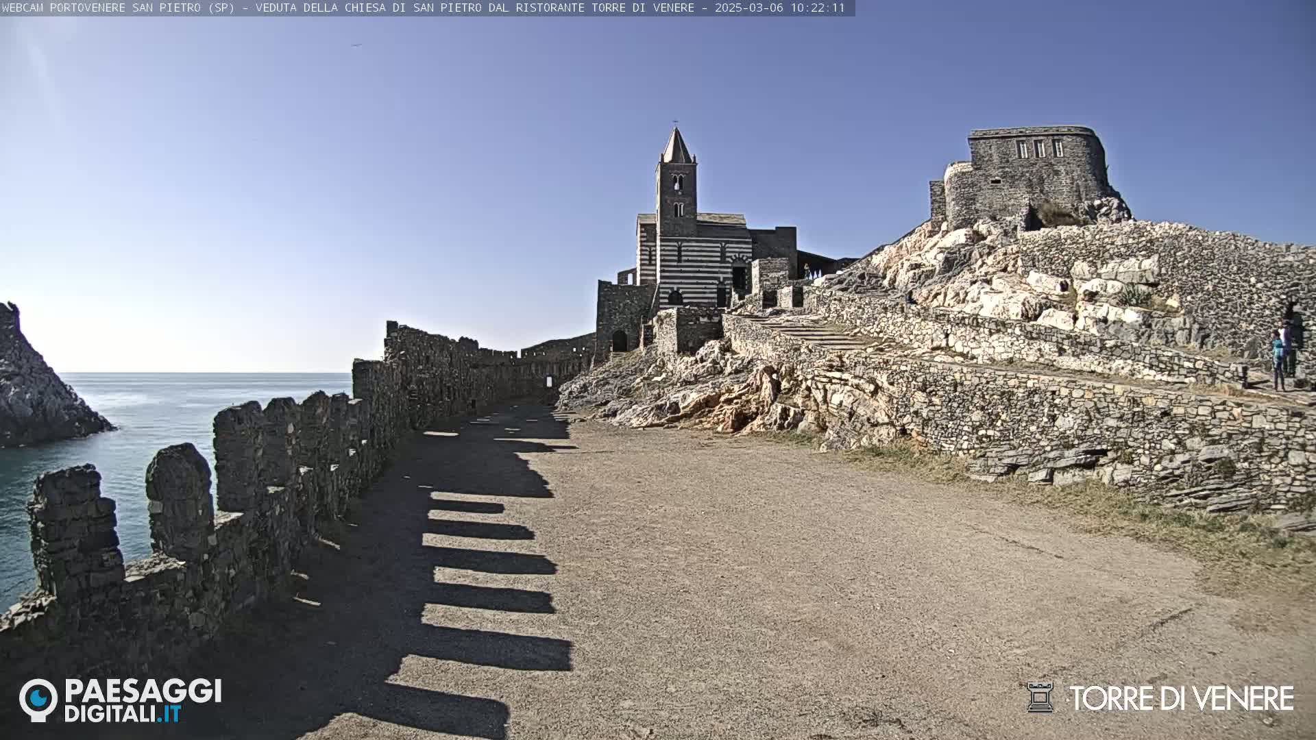A coastal stone pathway leads to a striped church and castle ruins under a clear blue sky.