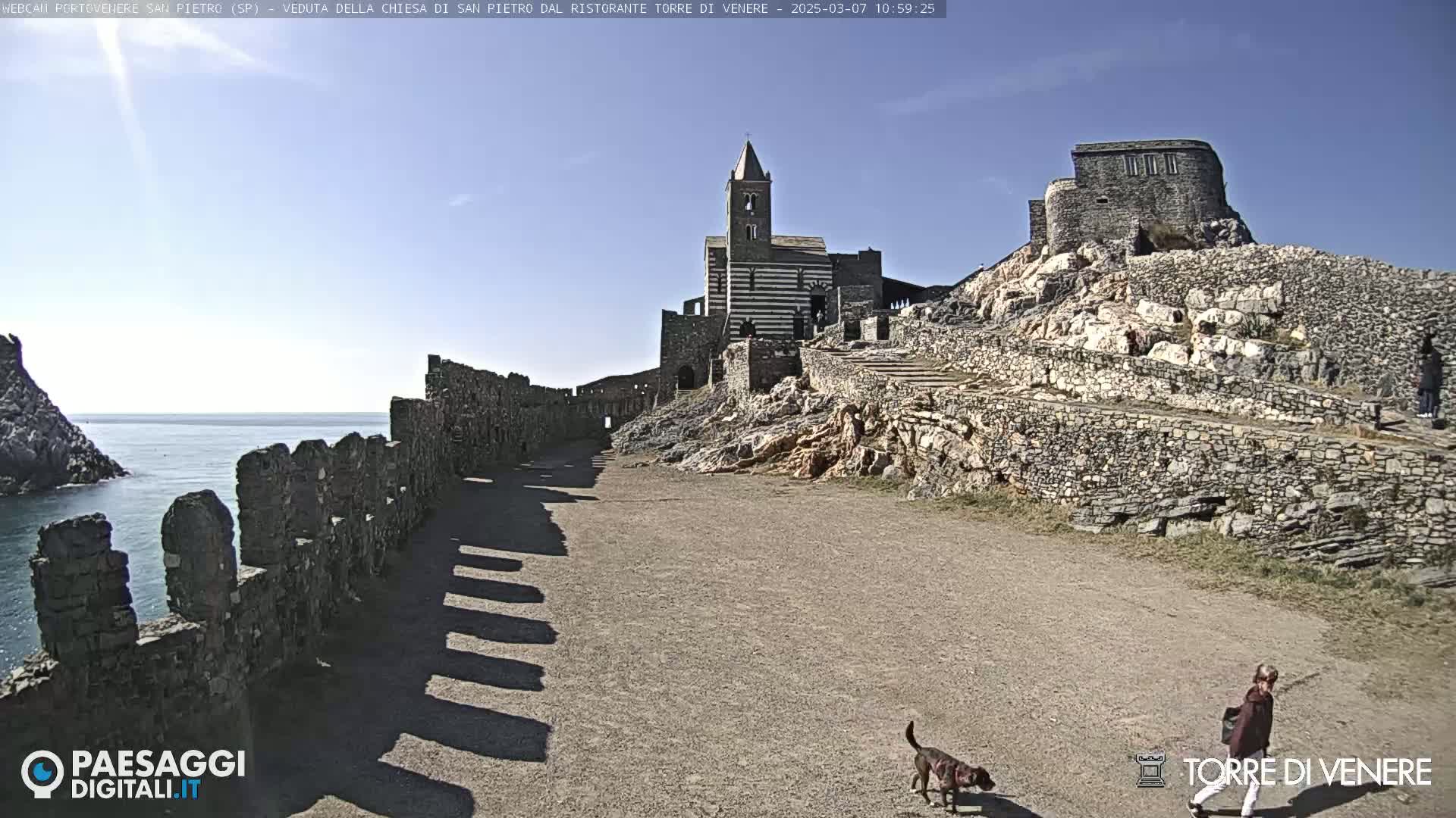 A stone pathway leads toward a striped church and castle ruins on a sunny day, with a person and dog walking along the path and the ocean visible in the background.